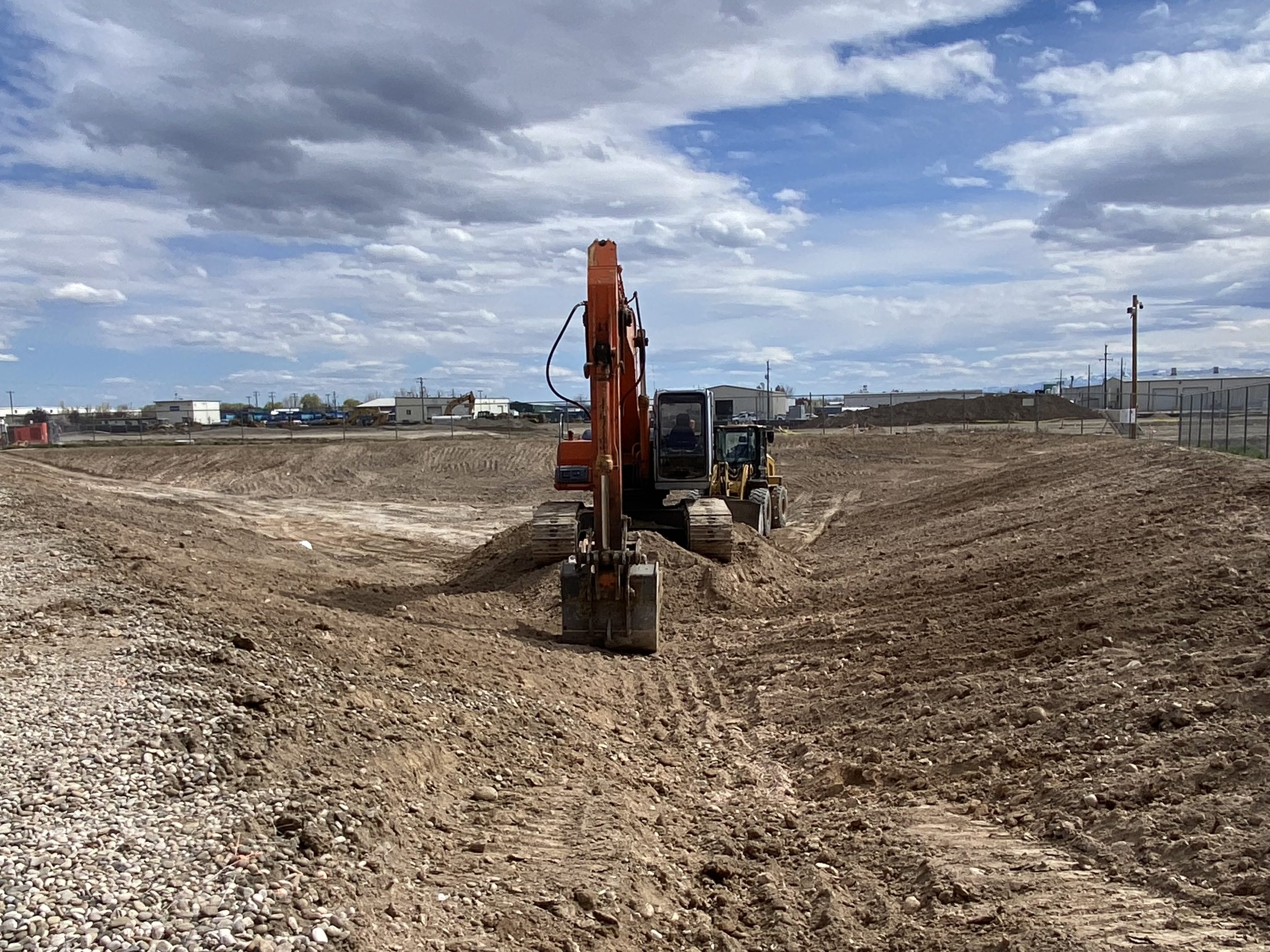 Excavator digging a large new retention pond with industrial buildings in the background